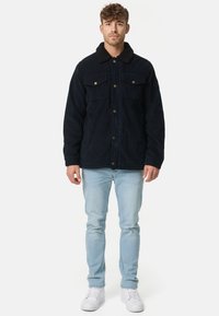 Marineblauwe corduroyjas met een zwarte bontkraag, twee borstzakken, messing knopen en een ontspannen pasvorm, gecombineerd met lichtblauwe jeans en witte sneakers.