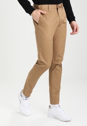 Person iført beige slim fit bukser med hånd i lommen, sort langærmet top, hvide sokker og hvide Reebok sneakers, der går.