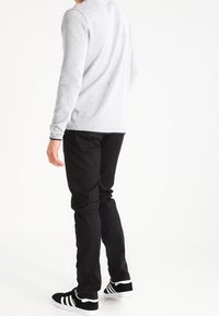 Šedý svetr s dlouhým rukávem, černé slim-fit kalhoty a černé sportovní boty s bílými pruhy. Tkanina vypadá strukturovaně a měkce.