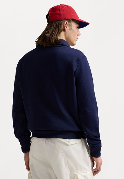Mørkeblå sweatshirt med lange ærmer, lavet af et blødt materiale. Parret med en rød kasket, set fra bagen, med minimal detaljer.