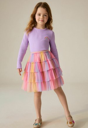 Fată tânără cu păr lung, purtând un top violet cu un patch în formă de curcubeu, o fustă în straturi multicolore și pantofi sclipitori cu modele de stele.