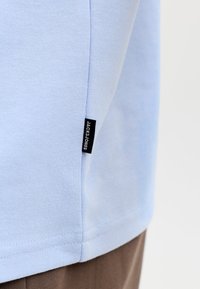 Šviesiai mėlyna medžiaga su siūlomu juodu „JACK & JONES“ žyme netoli siūlės, dėvima ant rudų kelnių.