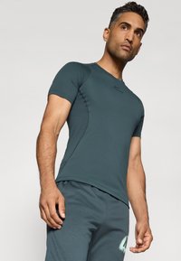 Homme portant un t-shirt de sport à manches courtes ajusté de couleur bleu sarcelle foncé et un short assorti, se tenant debout et regardant légèrement vers le haut sur un fond uni.