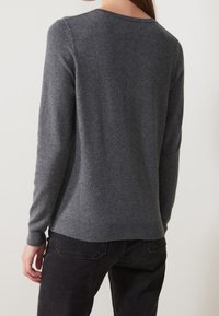 Kobieta w dopasowanej, ciemnoszarej, długiej bluzie z długim rękawem i czarnych dżinsach, pokazana od tyłu na tle jasnym.