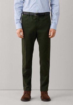 Man wearing dark green trousers, brown suede shoes, black belt, and light blue striped long-sleeve shirt standing against a plain background.
