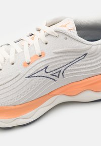 Chaussure de sport gris clair avec un dessus en tricot texturé, des coutures d'accent en bleu marine et une semelle intermédiaire en caoutchouc orange. Présente des lacets traditionnels et une marque.