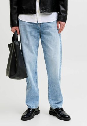 Personne portant un jean bleu clair, des chaussures en cuir noires, une chemise blanche et une veste en cuir noire tenant un sac fourre-tout en cuir noir.