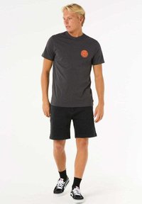 Graues Baumwoll-T-Shirt mit einem runden orangefarbenen Logo auf der linken Brust, kombiniert mit schwarzen Jeansshorts und schwarzen Sneakern mit weißen Sohlen.