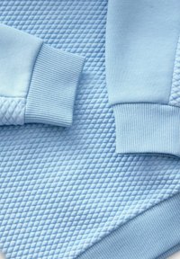 Jasnoniebieski sweatshirt z pikowanym wzorem i ściągaczami. Miękka tkanina podkreśla geometryczny wzór na powierzchni.