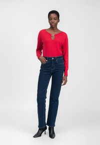Top rojo de manga larga con un detalle de ojal y acentos metálicos, combinado con jeans rectos de pierna oscura y botas de tobillo negras con tacón.
