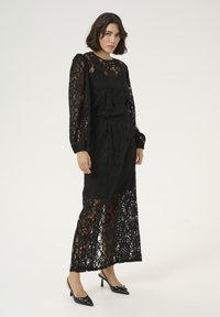 Robe en dentelle noire avec des manches longues, un design superposé et une texture semi-transparente. Associée à des escarpins noir à bride arrière, mettant en valeur des motifs délicats.
