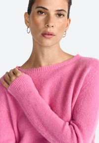 Maglione rosa lavorato a maglia con scollatura ampia e polsini a costine. Il materiale appare morbido con una consistenza leggermente pelosa.