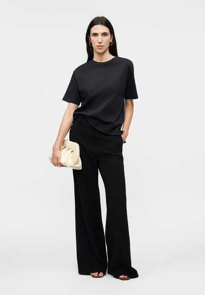 Γυναίκα με μακριά σκούρα μαλλιά, που φοράει μαύρο φαρδύ μπλουζάκι και παντελόνι με φαρδιά πόδια, κρατώντας μια μπεζ τσάντα φάκελο σε απλό ανοιχτό φόντο.