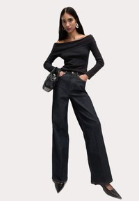 Zwarte off-shoulder lange mouwen top gecombineerd met hoge taille donkere denim wijde broek. Model houdt een zwarte handtas vast; puntige zwarte hakken maken de look af.