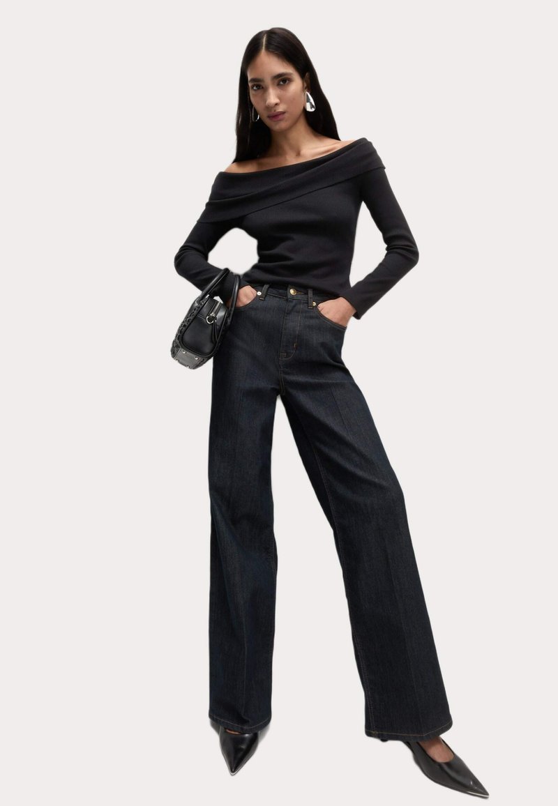 Zwarte off-shoulder lange mouwen top gecombineerd met hoge taille donkere denim wijde broek. Model houdt een zwarte handtas vast; puntige zwarte hakken maken de look af.