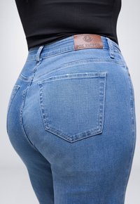 Close-up van een persoon die lichtblauwe denim jeans draagt met een bruine "Fox Factor" label op de tailleband en een zwarte top die erin is gestoken.