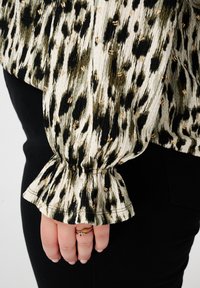 Blouse texturée en tissu à motifs crème, vert et noir, avec des manches évasées et de subtils accents dorés. Associée à un pantalon noir.