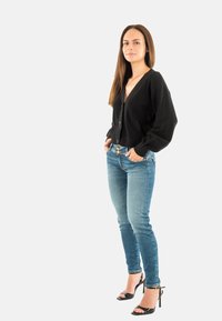 Cardigan nero corto con scollo a V, abbinato a jeans aderenti blu e tacchi alti neri aperti, che evidenziano un design elegante e una texture liscia.
