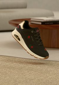 Baskets Skechers noires avec broderie de rose rouge et détail doré au talon sur un tapis beige près d'une table en bois avec des livres empilés.