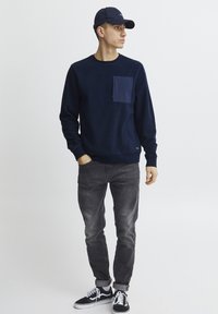 Mornarsko modra flis majica s sprednjim žepom, v kombinaciji z sivimi džinsom in črnimi copati z belimi podplati. Model nosi ujemajočo se kapo.