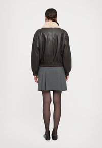 Woman standing with back to camera, wearing dark brown leather jacket with beige collar, gray pleated skirt, black tights, and black flats.