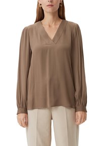 Blouse marron à manches longues avec un col en V, en tissu lisse, dotée de poignets élastiques. Portée avec un pantalon plissé de couleur claire.