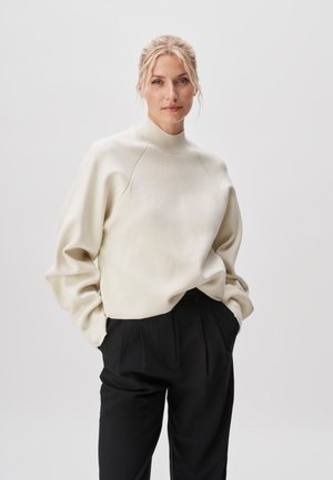 Femme blonde aux cheveux attachés portant un pull crème à col montant et un pantalon plissé noir, mains dans les poches, fond neutre.