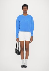 Μπλέ φούτερ με κείμενο "ΚΛΕΙΣΤΟ," λευκές βερμούδες, μαύρη τσάντα, λευκές κάλτσες και μαύρες φλατ εσπαντρίγιες. Το ύφασμα είναι απαλό, σε κανονική εφαρμογή.