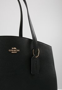 Sac fourre-tout Coach en cuir noir texturé avec doubles poignées, emblème logo doré et étiquette en cuir noir attachée à une chaîne à billes dorée.