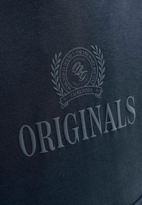 Emblème en relief avec une couronne de laurier et un texte circulaire au-dessus du mot « ORIGINALS » sur un tissu texturé bleu foncé.