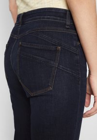 Tmavě modré džíny z denimu s úzkým střihem. Mezi vlastnosti patří dvě zadní kapsy, viditelné švy a hladká textura.