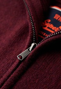 Nahaufnahme eines weinroten Wolljackenreißverschlusses mit schwarzen Metallzähnen und einem teilweise sichtbaren marineblauen und orangefarbenen Superdry-Label im Inneren.