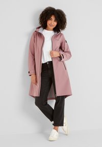 Pink waterproof jacket with a front zip, hood, and black accents. Worn over a white t-shirt and black jeans, paired with white sneakers.