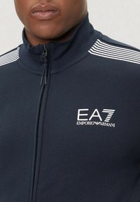 Mörkblå zip-up jacka med hög krage, vit logotyp och varumärkesinformation. Har randiga detaljer på axlarna och texturerad tyg.