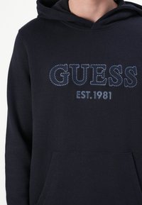 Felpa blu navy realizzata in morbido cotone con tasca a marsupio. Presenta "GUESS" e "EST.1981" in strass blu sul davanti.