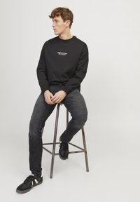 Sudadera negra con logo blanco, combinada con jeans oscuros y zapatillas negras, sentada en un taburete de metal frente a un fondo sencillo.