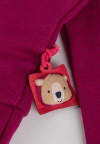 Fuchsia-Stoff mit glatter Textur zeigt ein rotes Etikett mit einem Bärenkopf-Design, das mit einem pinkfarbenen Kabel befestigt ist.