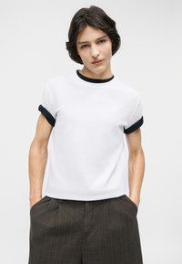 Νεαρός με κοντά σκούρα μαλλιά που φοράει λευκό μπλουζάκι με μαύρο περίγραμμα στον γιακά και τα μανίκια, με τα χέρια στις τσέπες σκούρων, φαρδιών παντελονιών.