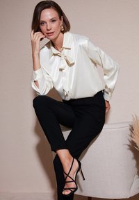 Blusa de satén crema con un lazo en el cuello, combinada con pantalones negros de corte ajustado y tacones negros con tiras. Fondo texturizado en tonos neutros.