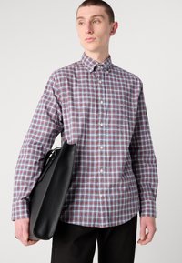 Camicia a maniche lunghe a quadri rossi e blu con colletto button-down, indossata con una borsa nera in pelle. Pantaloni neri semplici.