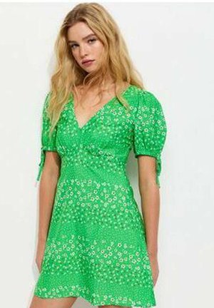 Vrouw met lang blond haar, gekleed in een groene korte jurk met bloemenprint en opgeblazen mouwen die bij de armen zijn gestrikt, staand tegen een lichte achtergrond.