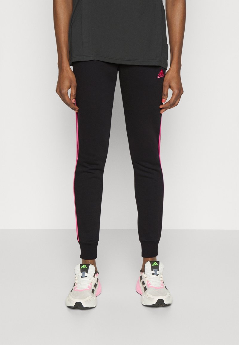 Person wearing black Adidas leggings with pink stripes and white Adidas sneakers with pink and green accents, standing against a white background.