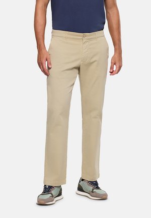 Hombre vistiendo pantalones beige de pierna recta, camiseta azul marino y zapatillas gris-verde, de pie contra un fondo liso.