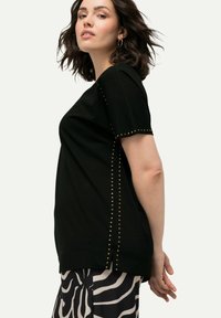 Camiseta negra de manga corta con tachuelas doradas a lo largo de las costuras laterales y los puños; tejido suave con un ajuste relajado. Se lleva con pantalones estampados.