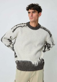 Jeune homme aux cheveux bouclés, portant un pull en peluche blanc et gris à motifs et un pantalon beige, debout avec les mains derrière le dos.