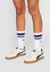 Krämfärgade sneakers med en svart rand och sula i gummi, kombinerade med vita strumpor med marinblå ränder och Puma-logotyp. Närbild av skorna.