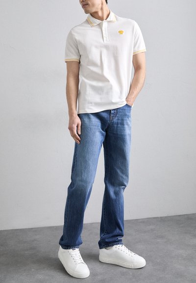 Cămașă polo albă cu guler în dungi galbene, având un logo. Purtați cu blugi din denim albastru și adidași albi pe un fundal gri neted.
