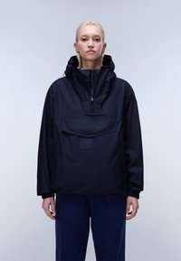 Γυναίκα με ξανθά μαλλιά φοράει μπλε σκούρο ανεμοφράκτη με κουκούλα, φερμουάρ μπροστά και μεγάλη τσέπη, στέκεται μπροστά σε απλό φωτεινό φόντο.