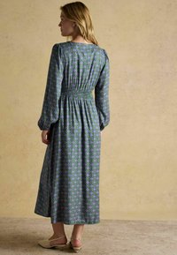 Dlhé, vzorované šaty v zelenej, purpurovej a modrej farbe so zúženým pásom a balónovými rukávmi. Doplněné béžovými topánkami.