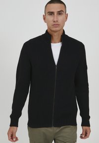 Dunkelblaue Zip-Up-Strickjacke mit hohem Kragen, gerippter Textur, langen Ärmeln und seitlichen Taschen. Über einem weißen Hemd getragen. Glatte Stoffoberfläche.